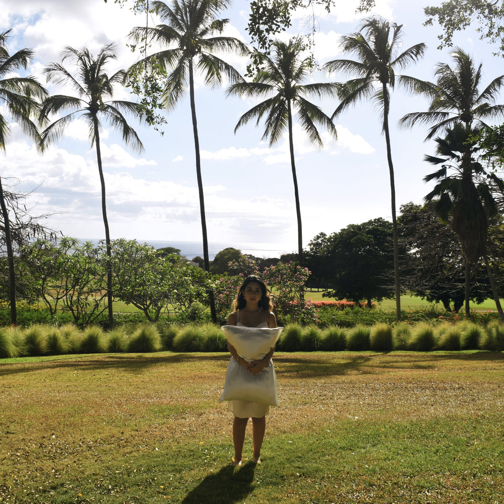 A woman stands on a tropical grassy lawn with tall palm trees, holding a white silk pillow.