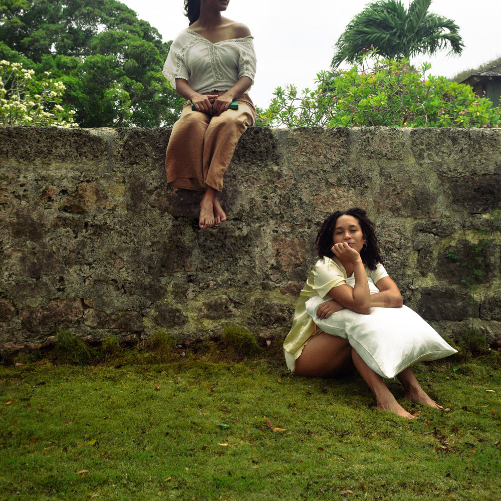 Two women in bohemian-style clothing pose outdoors against a stone wall with lush green foliage. One sits on the grass holding a white silk pillow, while the other is perched on the stone wall above.