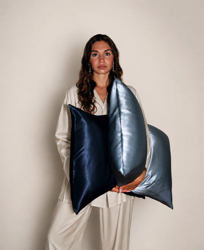 A woman in cream pajamas holds two large silk pillows in navy blue and gray blue against a plain beige studio background.