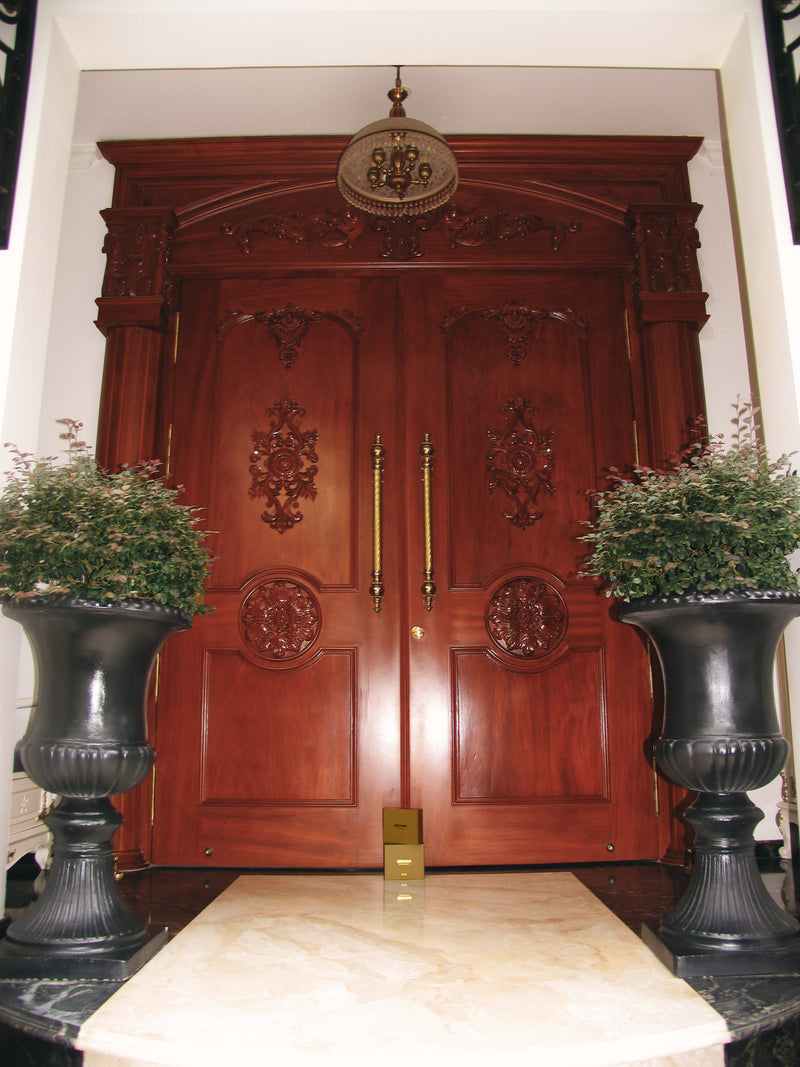 An ornate pair of dark reddish-brown wooden double doors with intricate carvings and gold-toned handles, set in a doorway with potted plants on either side and Evevior product boxes in front of the door.
