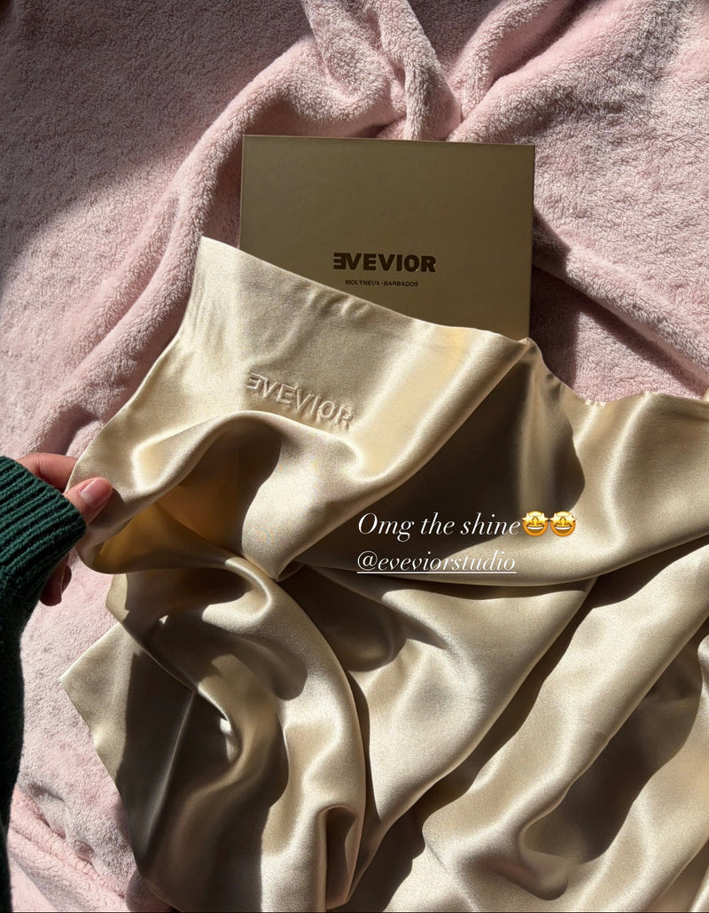 Hand holding a champagne silk pillowcase with embroidered Evevior branding, its sheen catching the sunlight against a pink blanket, with an Evevior box behind it. Text reads “Omg the shine 🤩🤩 @eveviorstudio.”