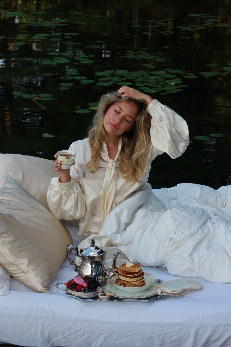 Woman in cream pyjamas sipping tea in bed on a lake, propped on champagne silk pillowcases with breakfast on a silver tray