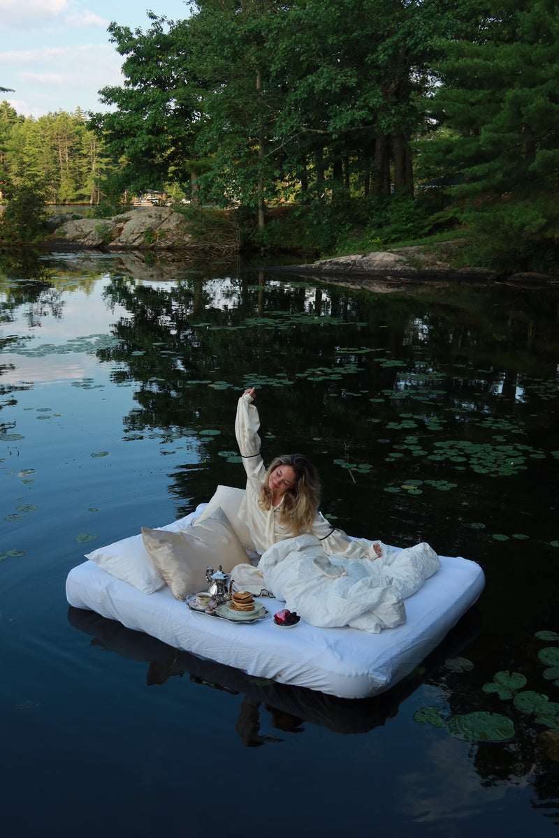 Woman stretching awake on a bed floating among lily pads on a forest lake, with champagne silk pillowcases and a breakfast tray