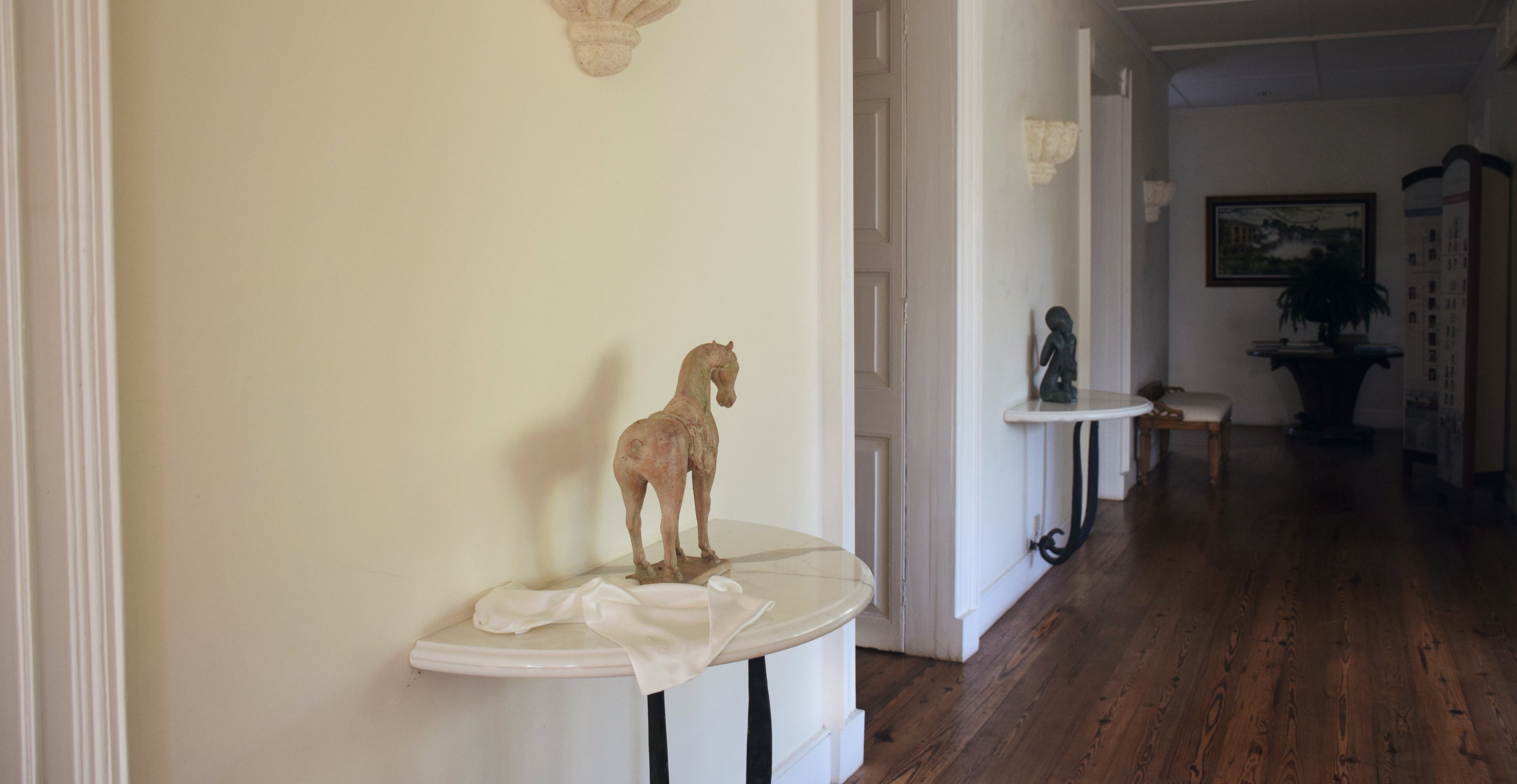 White silk pillowcase draped over a marble side table beside an antique horse sculpture, in a colonial-style hallway with dark wood floors.