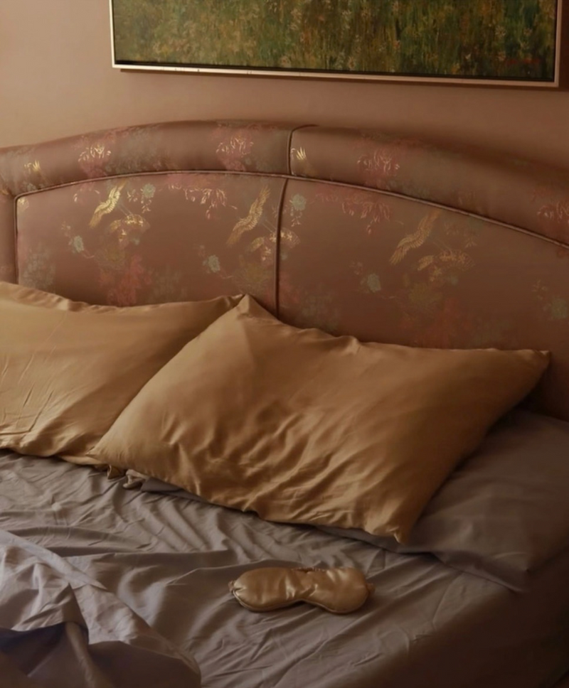 Two Nougat Evevior silk pillowcases and a silk eye mask on a bed with a floral upholstered headboard