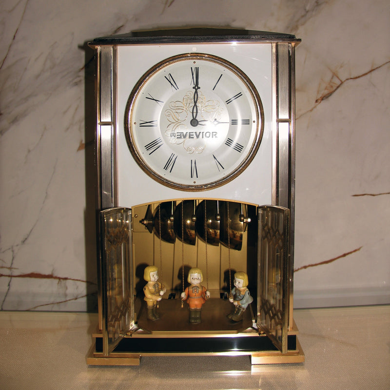 A vintage-style Kevior mantel clock with a polished gold-toned metal frame, white clock face, and three small figurines inside a glass-enclosed lower chamber, set against a marble-patterned background.