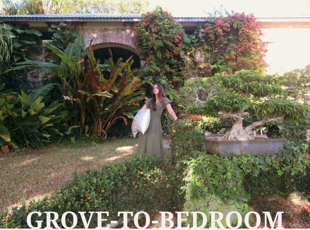 Woman in an olive green dress holding a white silk pillowcase in a lush tropical garden, surrounded by greenery, vibrant pink flowers and soft natural sunlight. Words on bottom of image read: GROVE-TO-BEDROOM