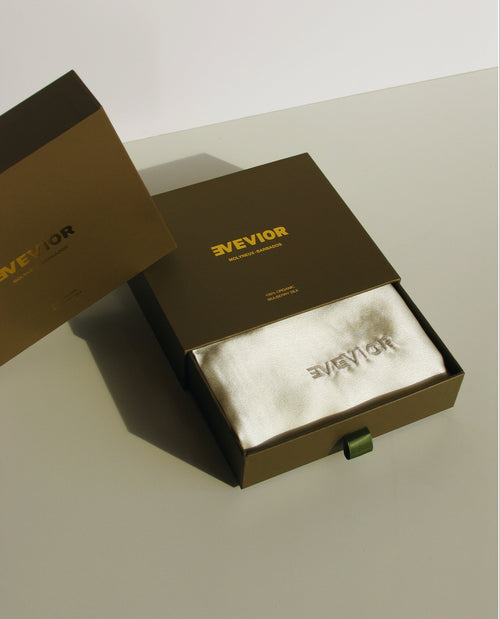A luxury product presentation showing a dark green "EVEVIOR" box with its compartment pulled out, revealing a cream silk pillowcase with "EVEVIOR" embroidered on it.