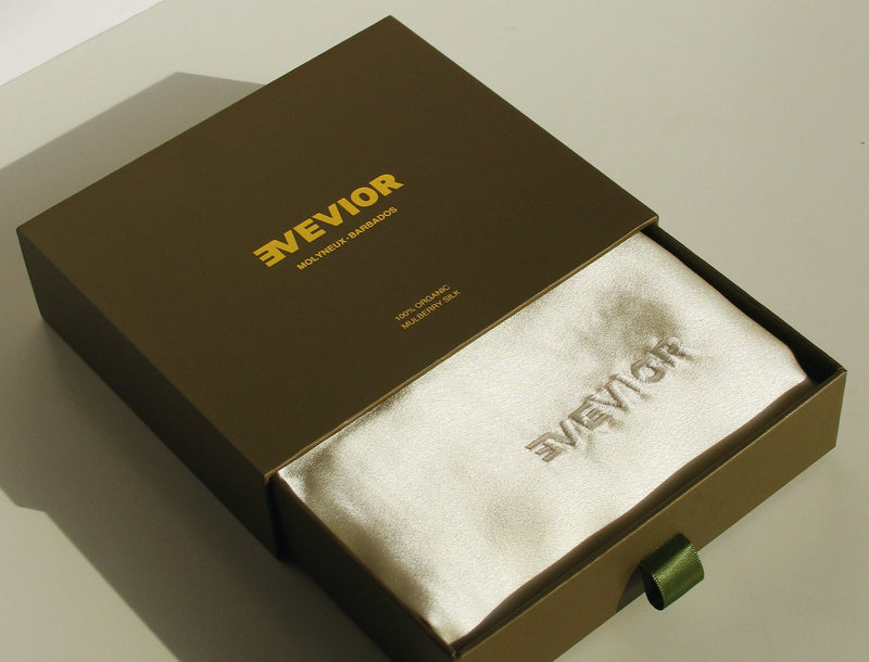 A luxury presentation box for EVEVIOR, featuring a dark green exterior with gold lettering and an open drawer containing a white silk pillowcase with the brand logo embroidered.