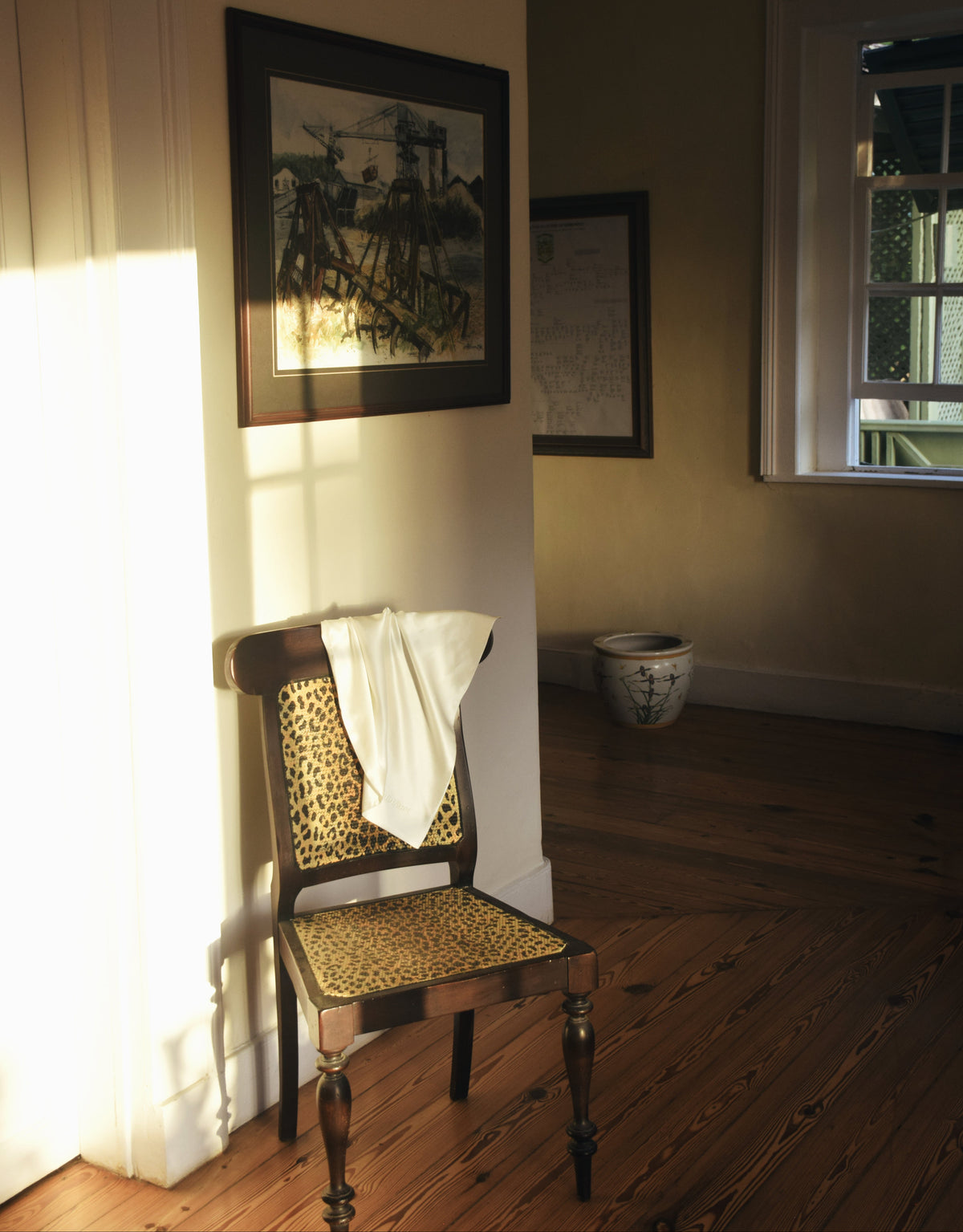 Leopard-print chair with a white silk pillowcase draped over the back, in a sunlit room with framed artwork and wood floors.