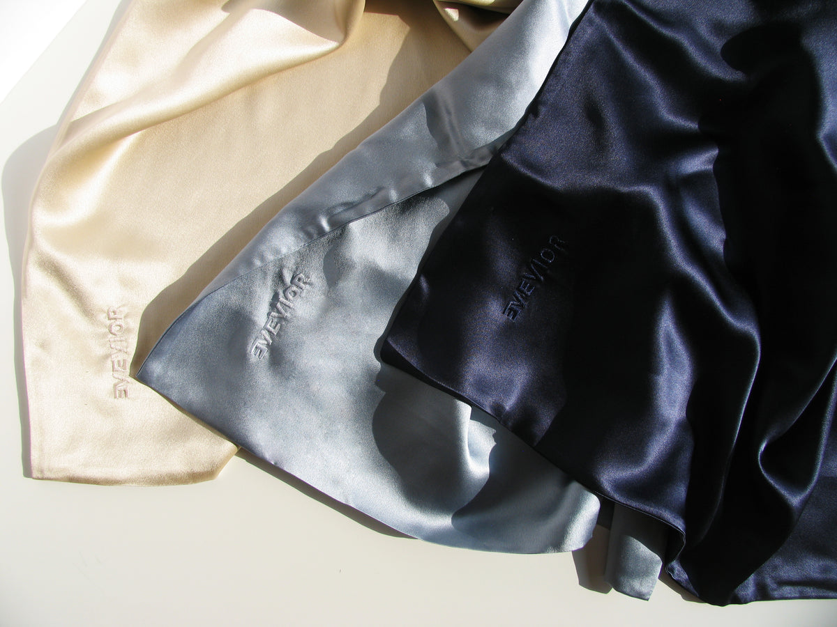 Three silk fabric pieces with 'EVEVIOR' embroidered on them, arranged diagonally. The fabrics are cream/beige, light blue-gray, and dark navy blue, shown under warm directional sunlight with soft shadows.