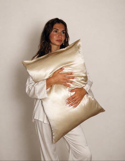 A woman in white pajamas holds a large, champagne colored silk pillow against her torso, posed against a plain light beige studio background.