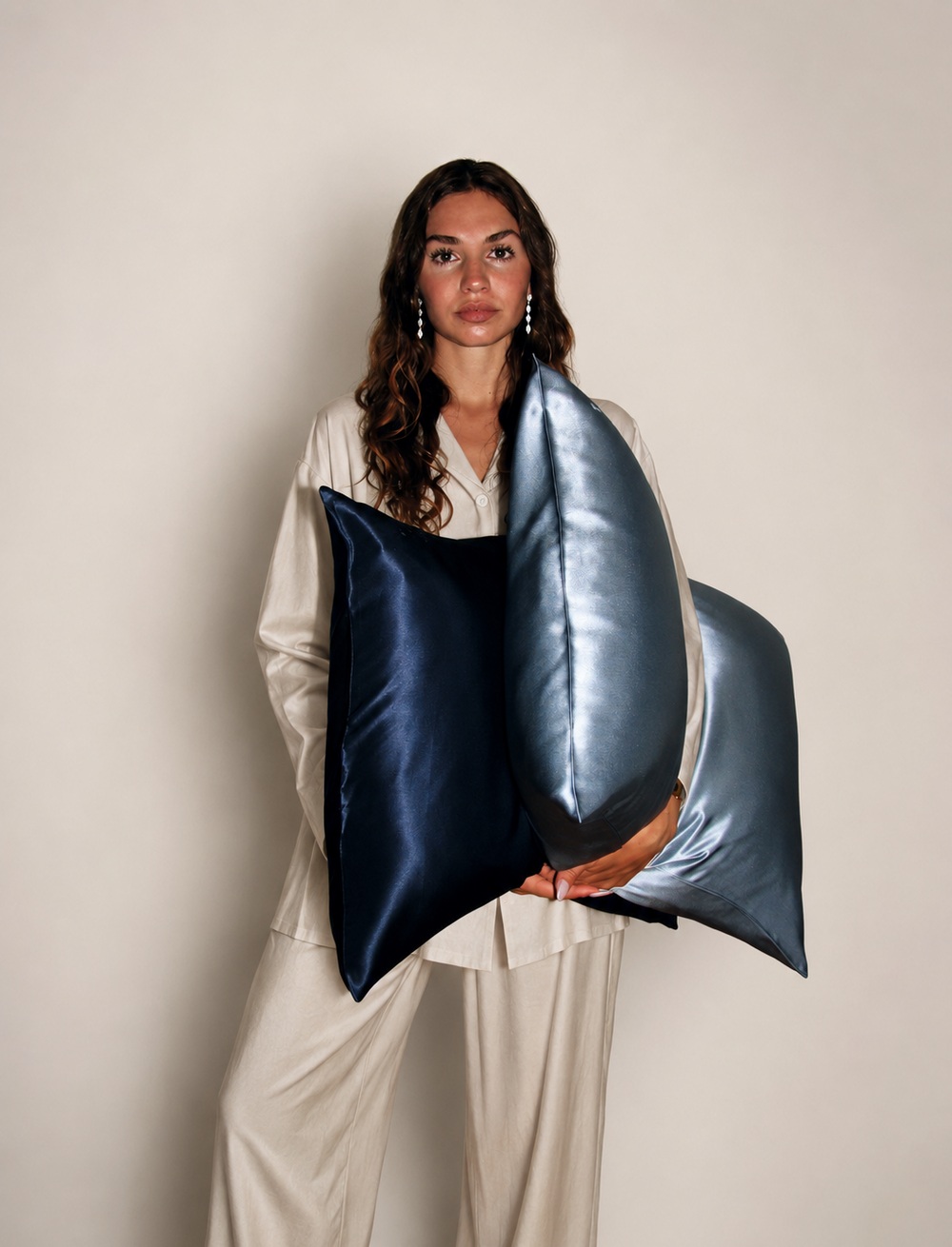 A woman in cream pajamas holds two large silk pillows in navy blue, grey blue  against a plain beige studio background. 