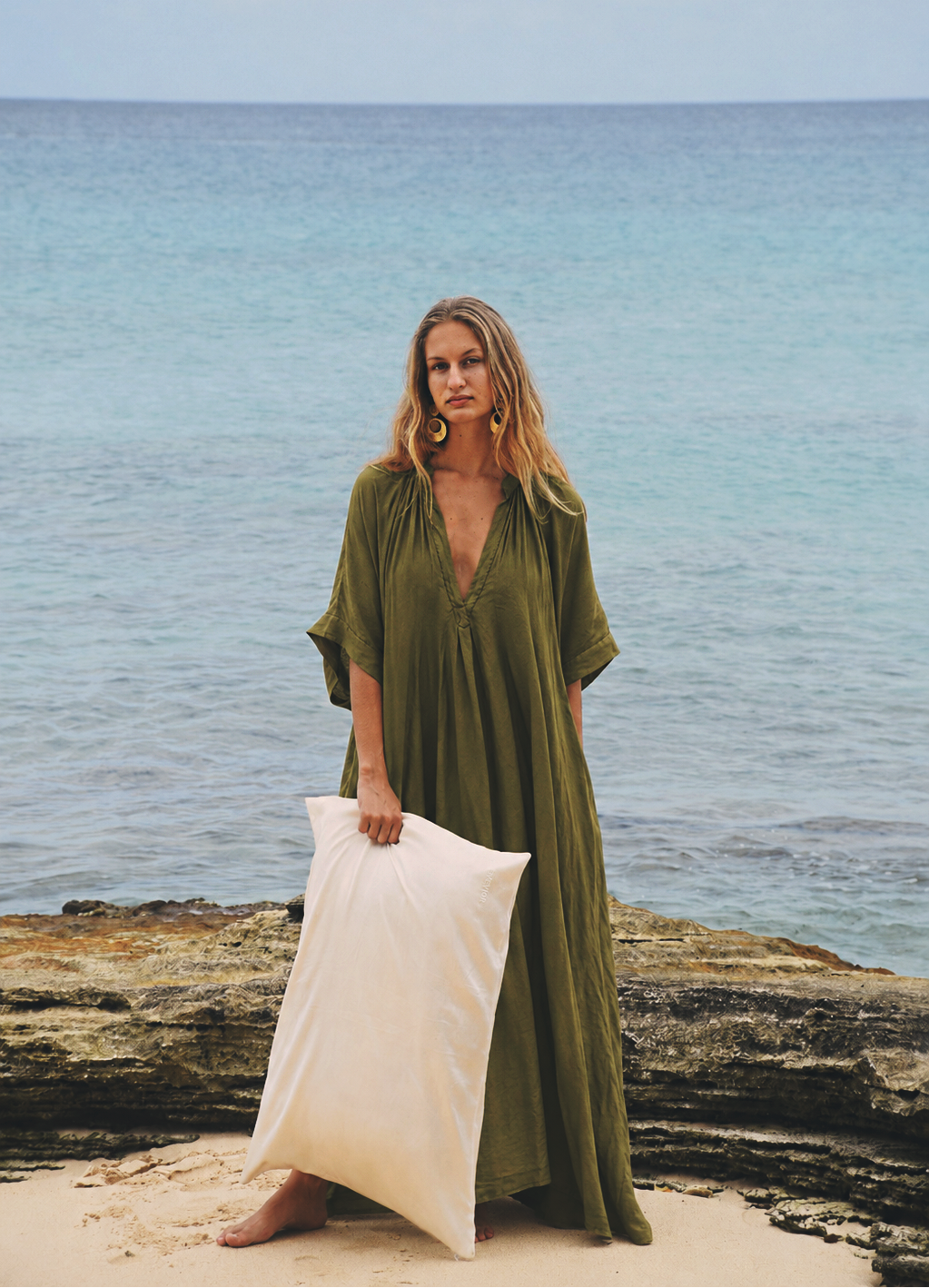 A woman stands on a sandy beach holding a large white silk pillow, wearing a loose olive green maxi dress and large gold hoop earrings, with the ocean and rocks behind her.