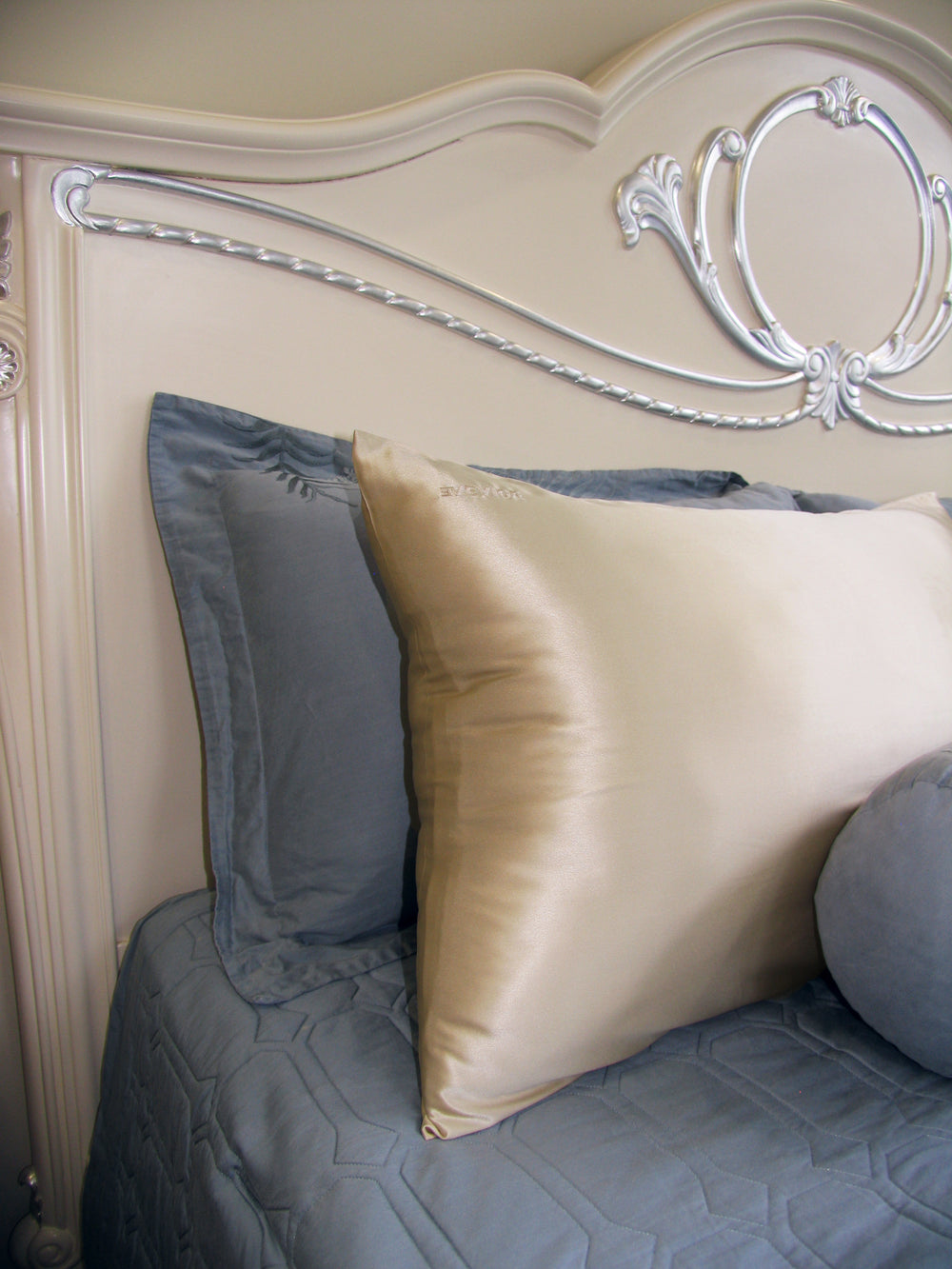A close-up of a bed with a decorative cream headboard and a grey quilted comforter, featuring a large cream silk pillow.