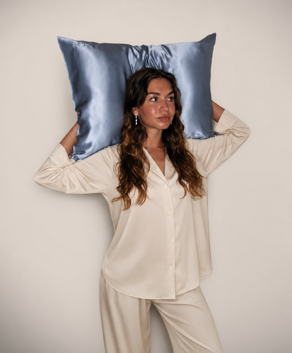 A woman wearing cream pajamas holds a grey blue silk pillow behind her head against a plain light beige backdrop.