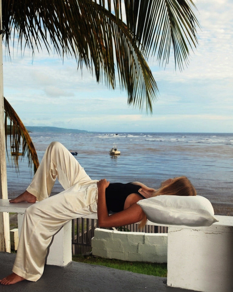 A woman in a black tank top and cream-colored silk pants lounges on a white outdoor bench with an ocean view in the background.