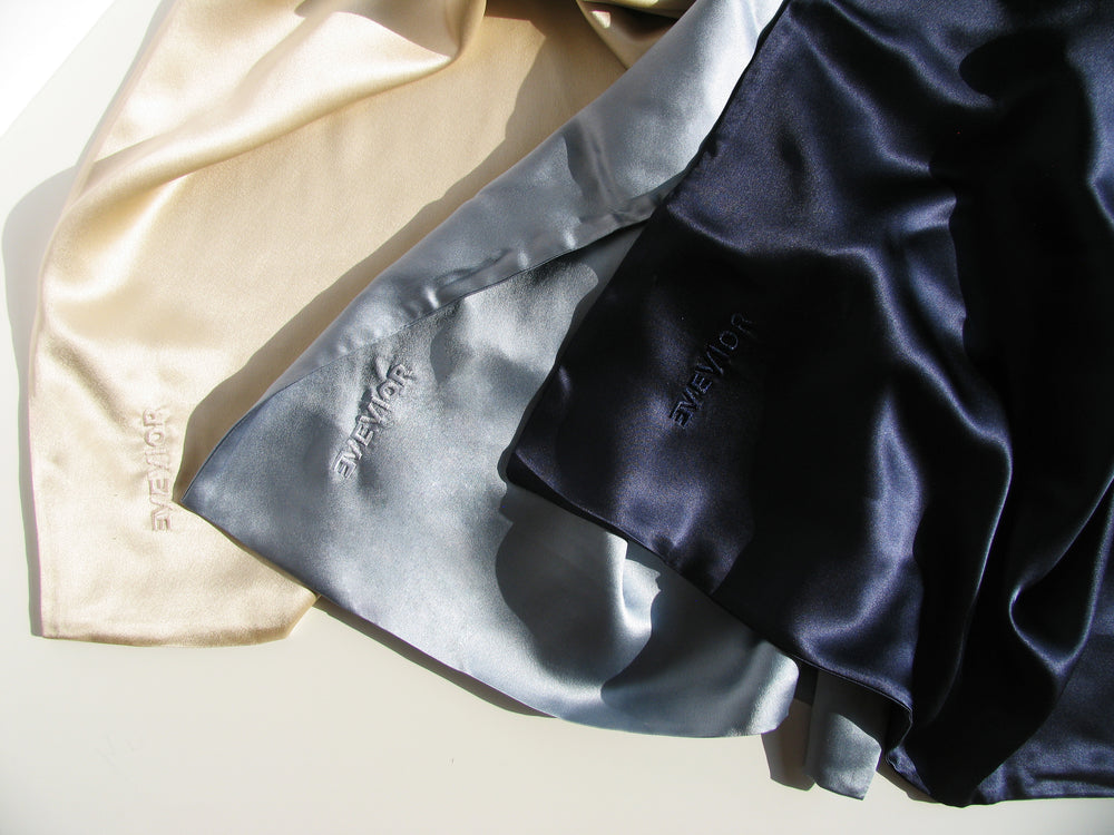 Three silk pillowcases pieces with 'EVEVIOR' embroidered on them, arranged diagonally. The fabrics are beige, light blue-gray, and dark navy blue, shown under warm directional sunlight with soft shadows.