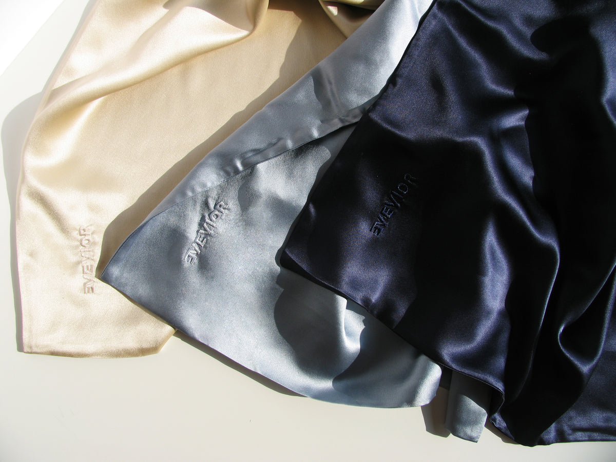 Three silk pillowcases pieces with 'EVEVIOR' embroidered on them, arranged diagonally. The fabrics are beige, light blue-gray, and dark navy blue, shown under warm directional sunlight with soft shadows.