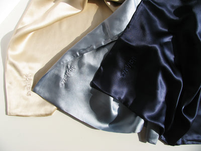 Three silk pillowcases pieces with 'EVEVIOR' embroidered on them, arranged diagonally. The fabrics are beige, light blue-gray, and dark navy blue, shown under warm directional sunlight with soft shadows.