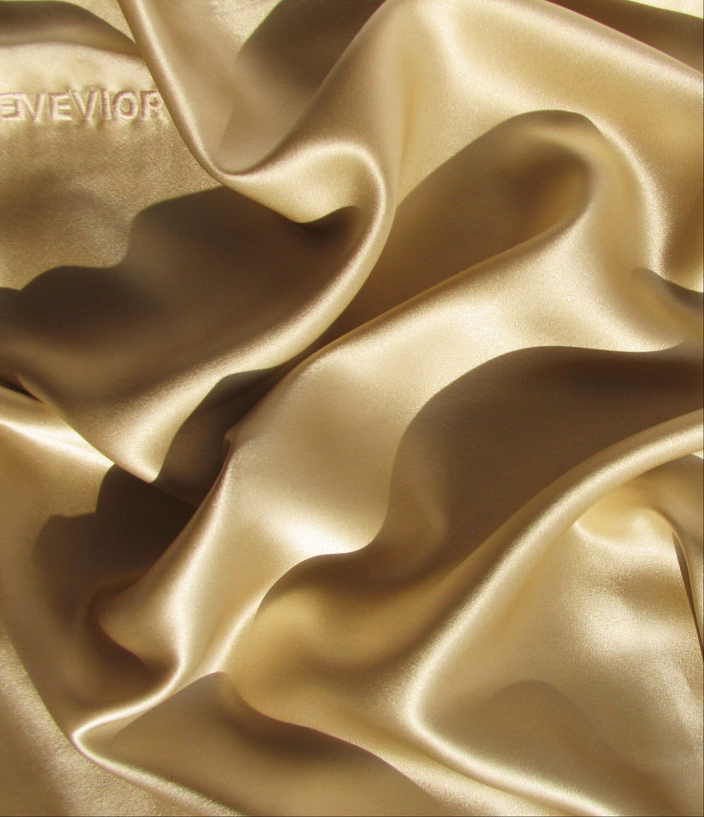 Close-up of draped champagne-gold silk pillowcase fabric with soft folds and a subtle sheen. Logo "EVEVIOR" is embroidered in the top left corner. 