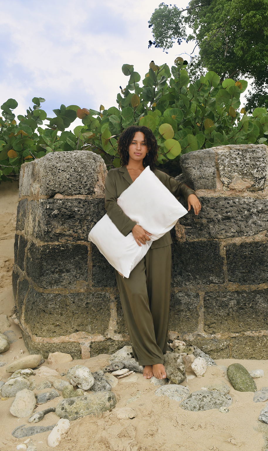 A woman stands barefoot on sandy ground in front of a stone structure, holding a large white silk pillow. She is wearing an olive green lounge set, with green plants in the background.