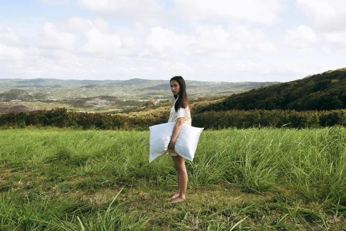 Woman holding a large white silk pillow in a grassy field with a scenic background.