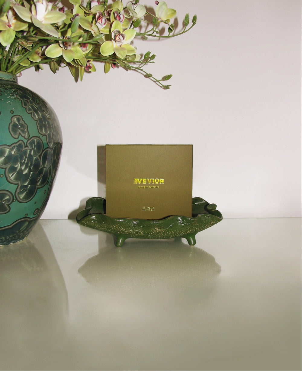 A square olive-green product box with gold lettering reading 'EVEVIOR' sits on a decorative green leaf-shaped tray, with a large green floral vase and pale flowers partially visible on the left against a light neutral background.