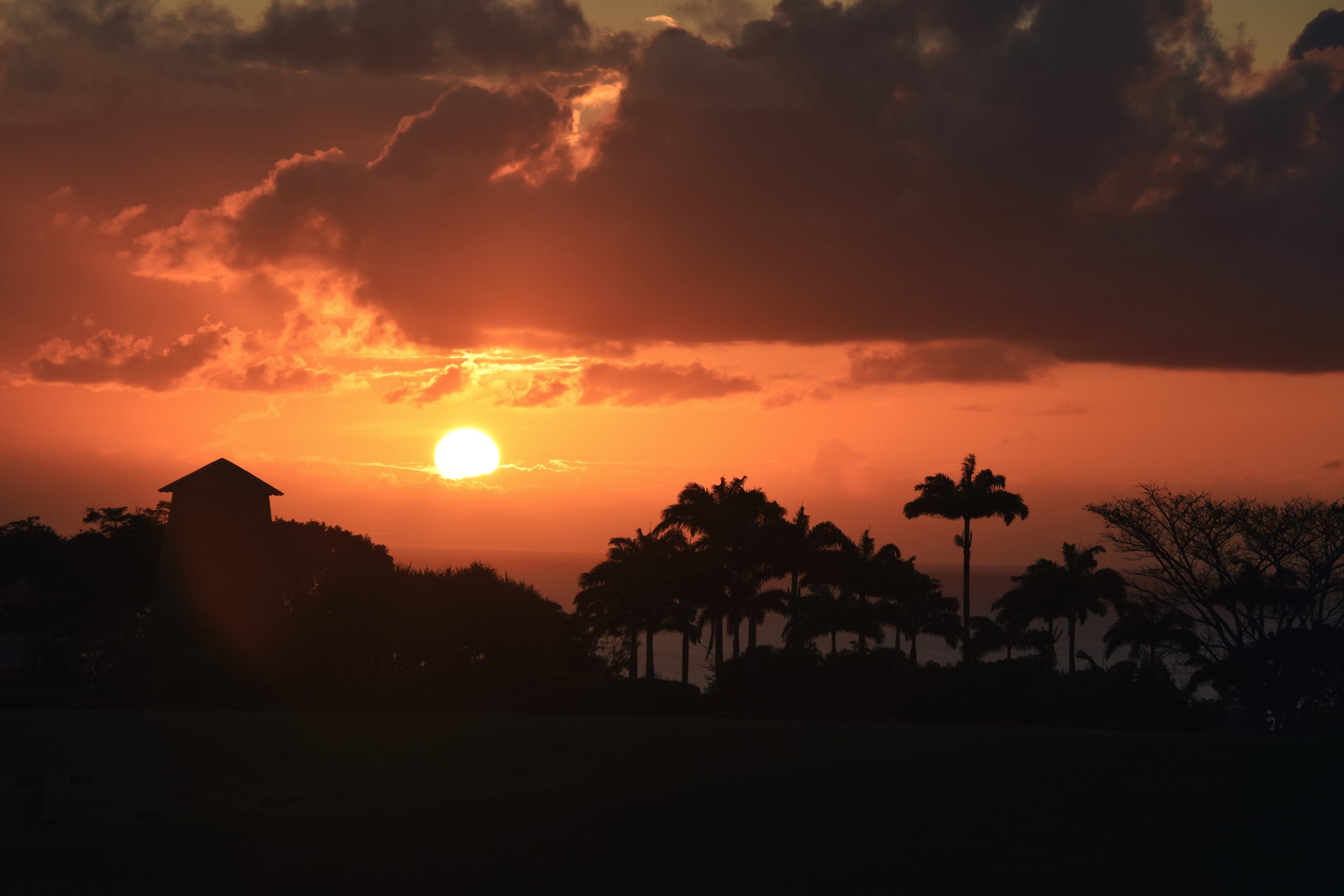 Sunset over a silhouette of trees and a sugar mill with clouds in the sky.